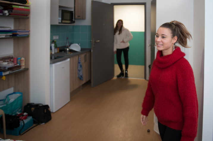 jeune femme souriant dans son appartement avec cuisine équipée. Une deuxième jeune femme est debout à l'entrée de l'appartement.